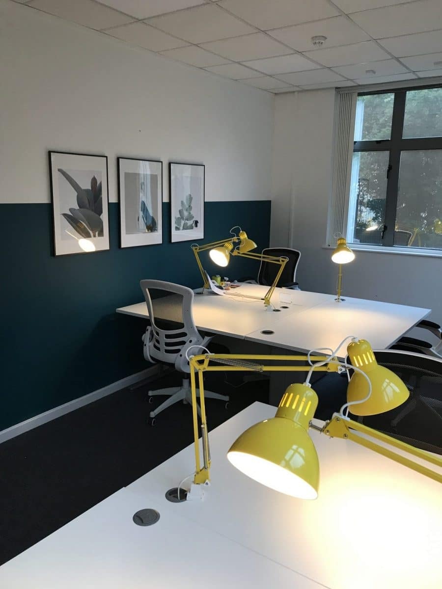 Small furnished office with two desks, yellow desk lamps and a teal wall